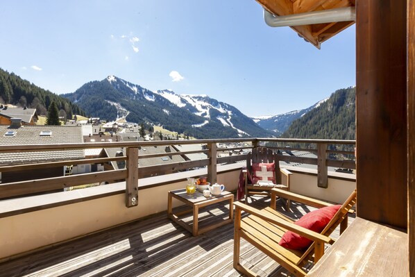 A balcony with wooden furniture, a table with a pitcher and teapot, and a view of snow-capped mountains.