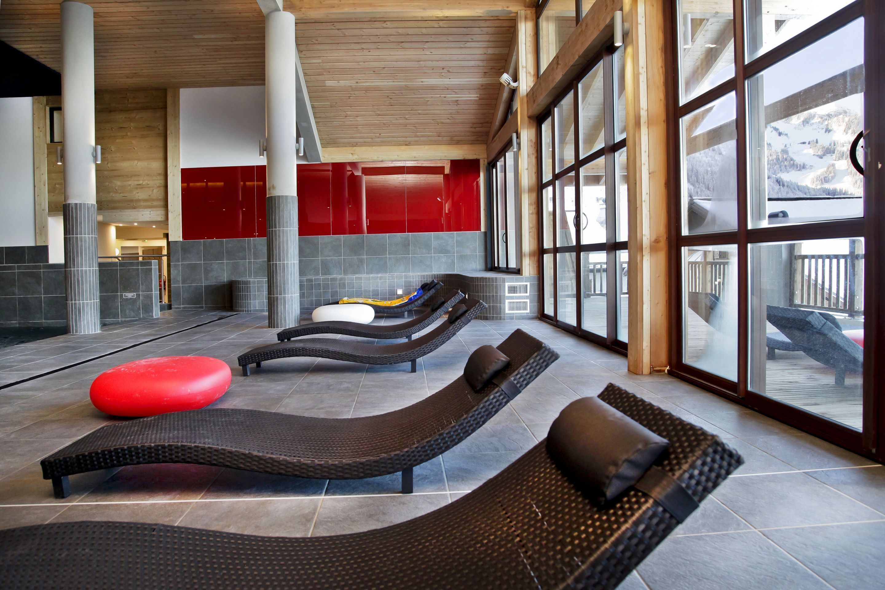 Un espace détente avec des chaises longues, un pouf rouge et de grandes fenêtres donnant sur l’extérieur.