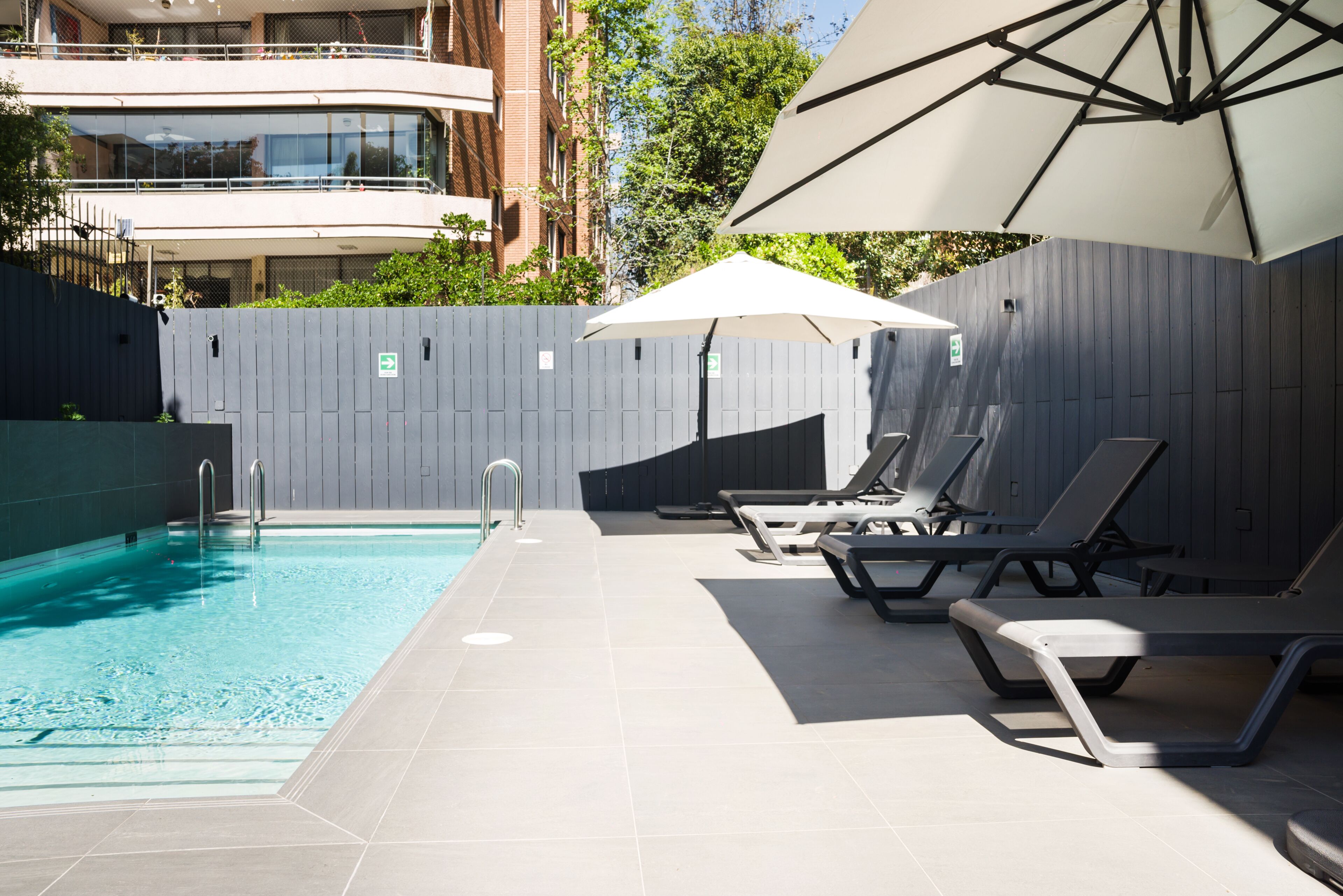 A swimming pool with lounge chairs and umbrellas, surrounded by a grey fence and a building in the background.