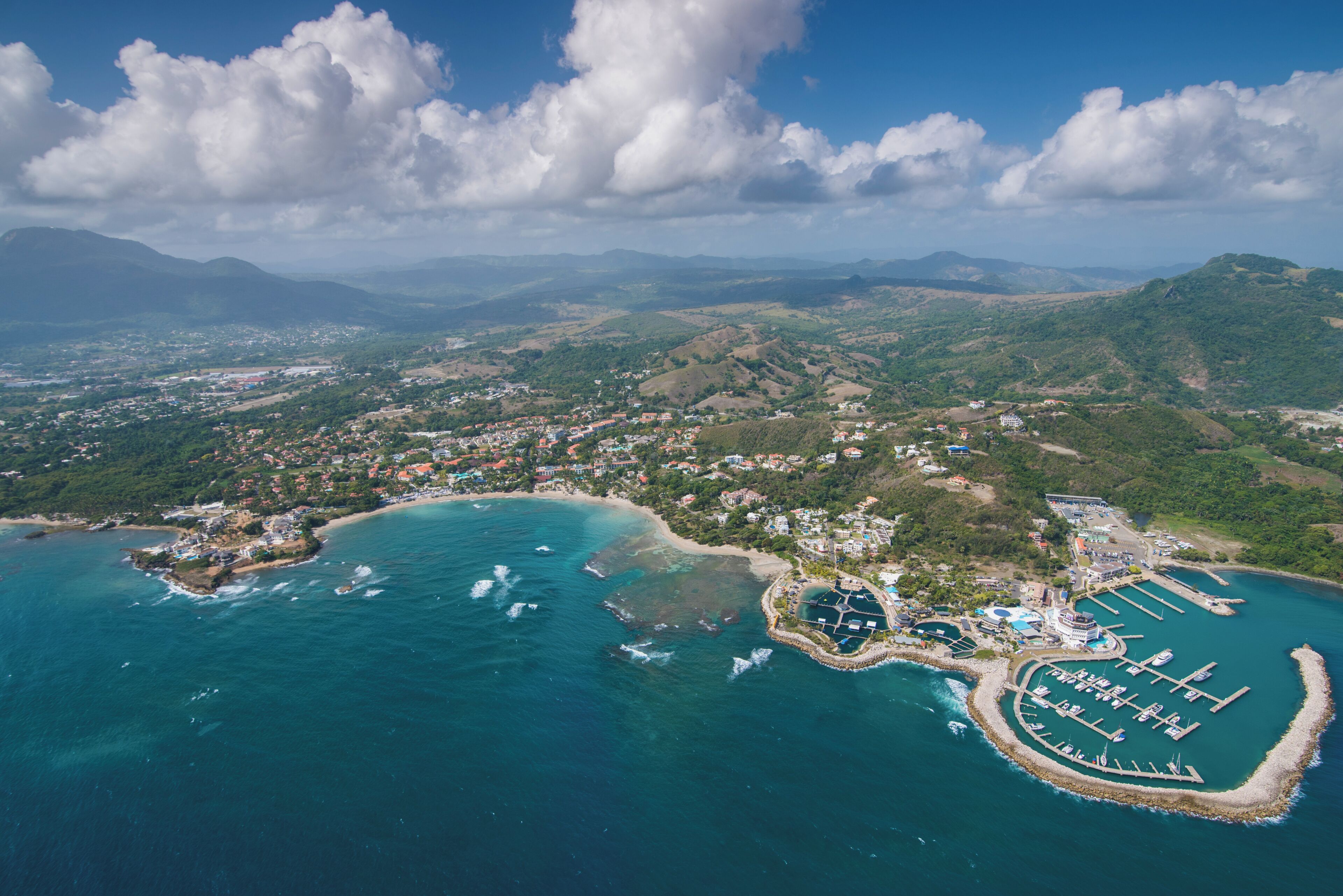 Vista aérea de un pueblo costero con un puerto deportivo, zonas residenciales y colinas circundantes.