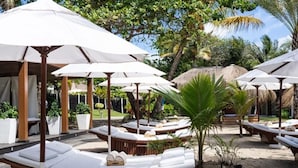 Gemütliche Lounge-Stühle mit weißen Kissen und Sonnenschirmen, in einer tropischen Umgebung mit Palmen und einer strohgedeckten Struktur im Hintergrund.