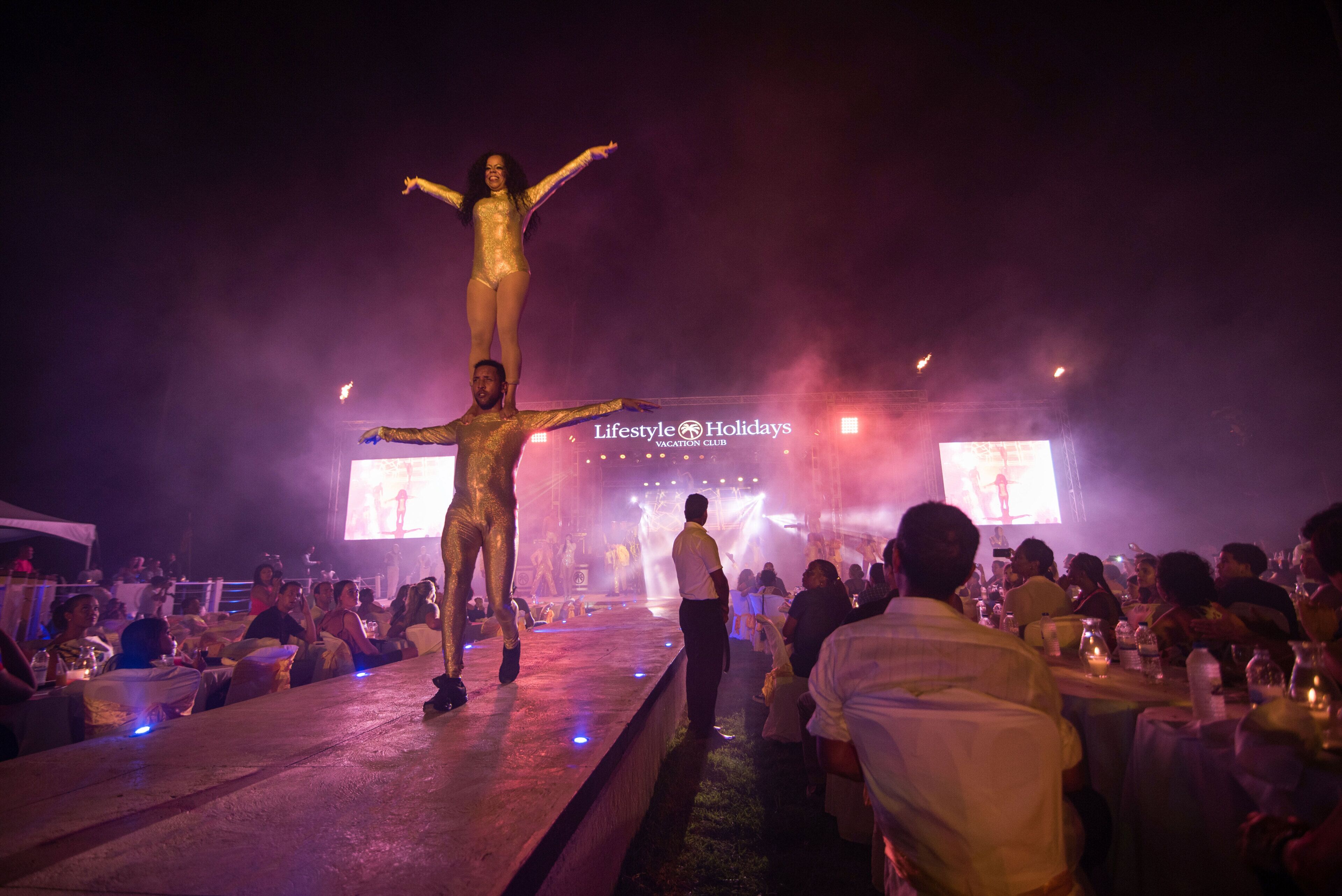 A stage with a performer in a golden costume, an audience seated at tables, and a large screen in the background.