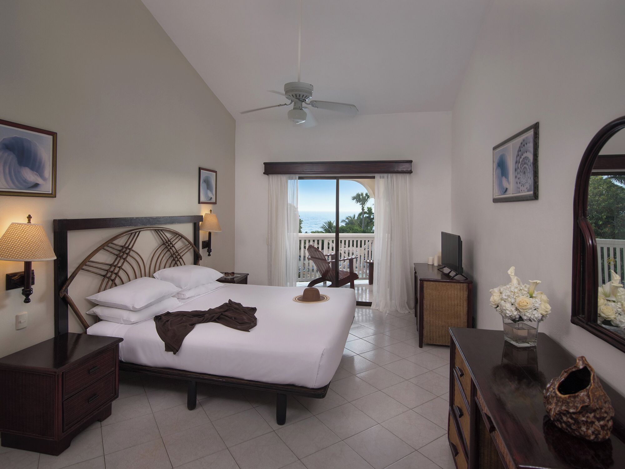 A hotel room with a bed, a nightstand, a dresser, a TV, a ceiling fan, and a balcony with a view of the sea.