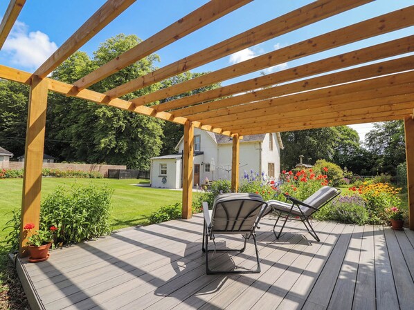 A wooden deck with two chairs, surrounded by a garden with various flowers and a house in the background.