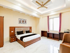 A bedroom with a bed, bedside tables, a desk, a chair, a ceiling fan, and a window with curtains. - Deluxe Kathauta Lake Formerly Shivanya (Lucknow)