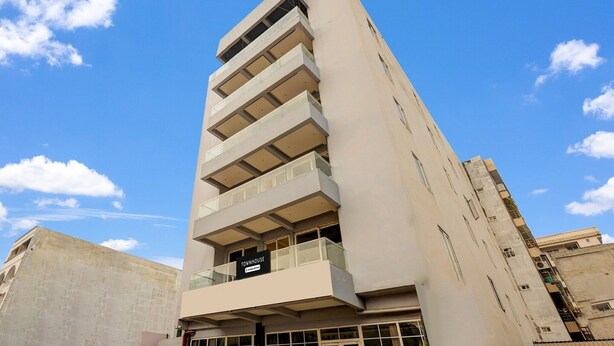 Un edificio de varios pisos con un balcón destacado en la planta baja, un letrero que indica que es una casa adosada, y un cielo azul despejado al fondo.