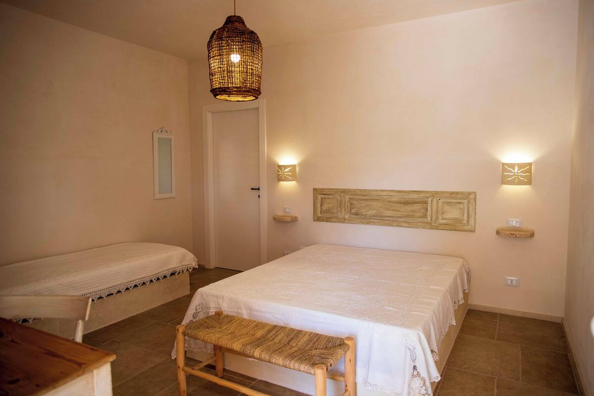 Quarto com cama, um banquinho, luminária pendente e iluminação na parede.