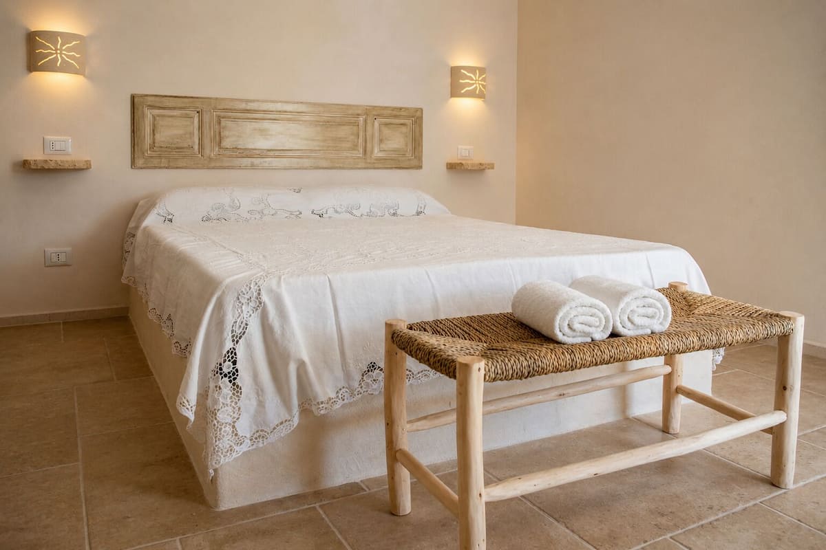 Cama bem arrumada com colcha branca e detalhes em renda, um banquinho de madeira com toalhas dobradas e duas luminárias fixadas na parede.