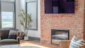 A living room with a brick wall, a fireplace, a flat-screen TV, and a leather sofa. - Stylish 3BR in Fountain Square Pool, Firepit, Bocce Court + Golden Tee (Indianapolis)