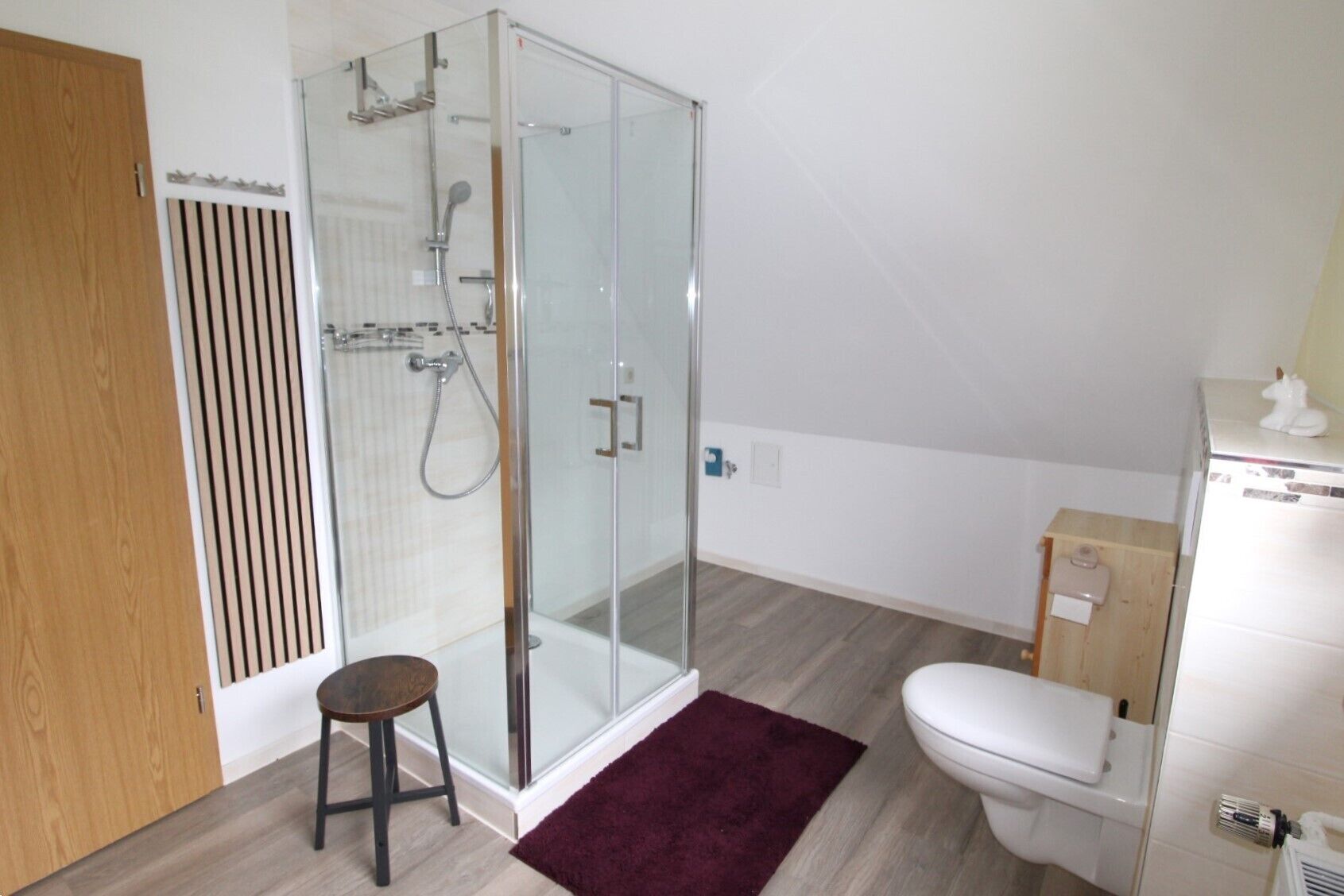A modern bathroom with a glass shower enclosure, a wooden door, a white toilet, and a small stool.