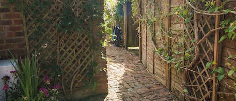 Un étroit sentier pavé de briques, bordé de clôtures en bois et de plantes grimpantes, mène à une porte en bois.