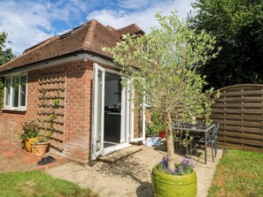 A brick house with a glass door, a small garden with a tree and potted plants, and a wooden fence in the background. - Lucerne Cottage (Dorking)