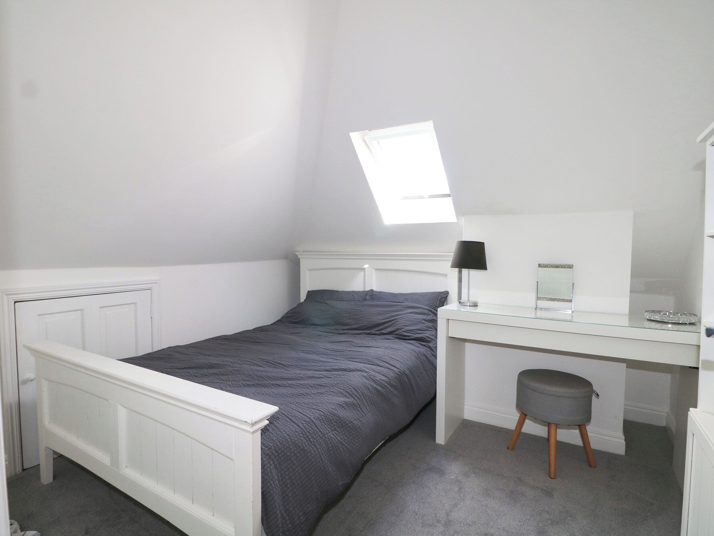 A bedroom with a bed, a desk, a chair, a small stool, a window, and a radiator.
