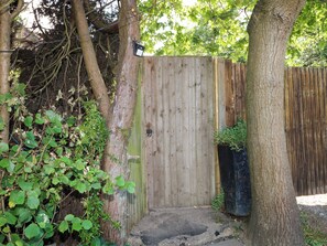 A wooden gate leading to a covered area with a small step, surrounded by trees and foliage. - Lucerne Cottage (Dorking)