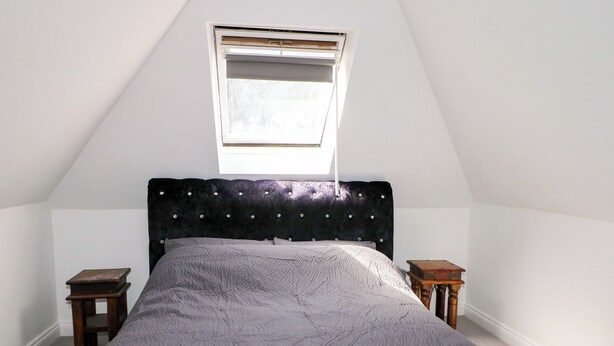 Un dormitorio con techo inclinado, una cama con cabecera capitonada, dos mesitas de noche y un ventanal.
