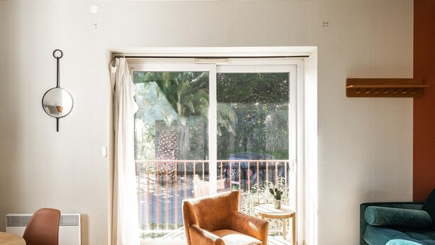 A living room with a green coffee table, a brown armchair, a blue sofa, a hanging light fixture, and a large sliding glass door leading to a balcony. - Studio Standard (Saint-Jean-de-Luz)