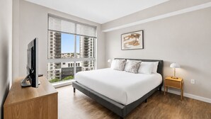 Een moderne slaapkamer met een groot bed, een tv op een houten bureau, een nachtkastje met een lamp, en uitzicht op de stad door ramen van de vloer tot aan het plafond.