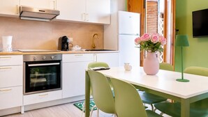 Une cuisine moderne avec des armoires blanches, un four encastré et une machine à café. On y trouve une table à manger avec des chaises, un vase de fleurs et un lampadaire vert.