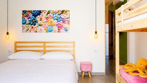 Une chambre avec un lit en bois, un petit tabouret rose, un lit superposé avec un jouet en peluche jaune, et un tableau coloré représentant des fleurs accroché au mur.