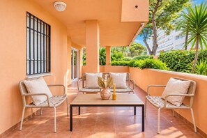 A covered outdoor seating area with wicker furniture and a central table, surrounded by a low wall and a view of greenery and buildings. - GuestReady - Marbella vibes with pool (Marbella)
