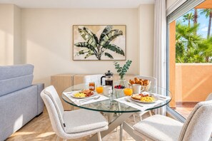 A dining area with a glass table, white chairs, breakfast items, and a large window with a view of palm trees. - GuestReady - Marbella vibes with pool (Marbella)