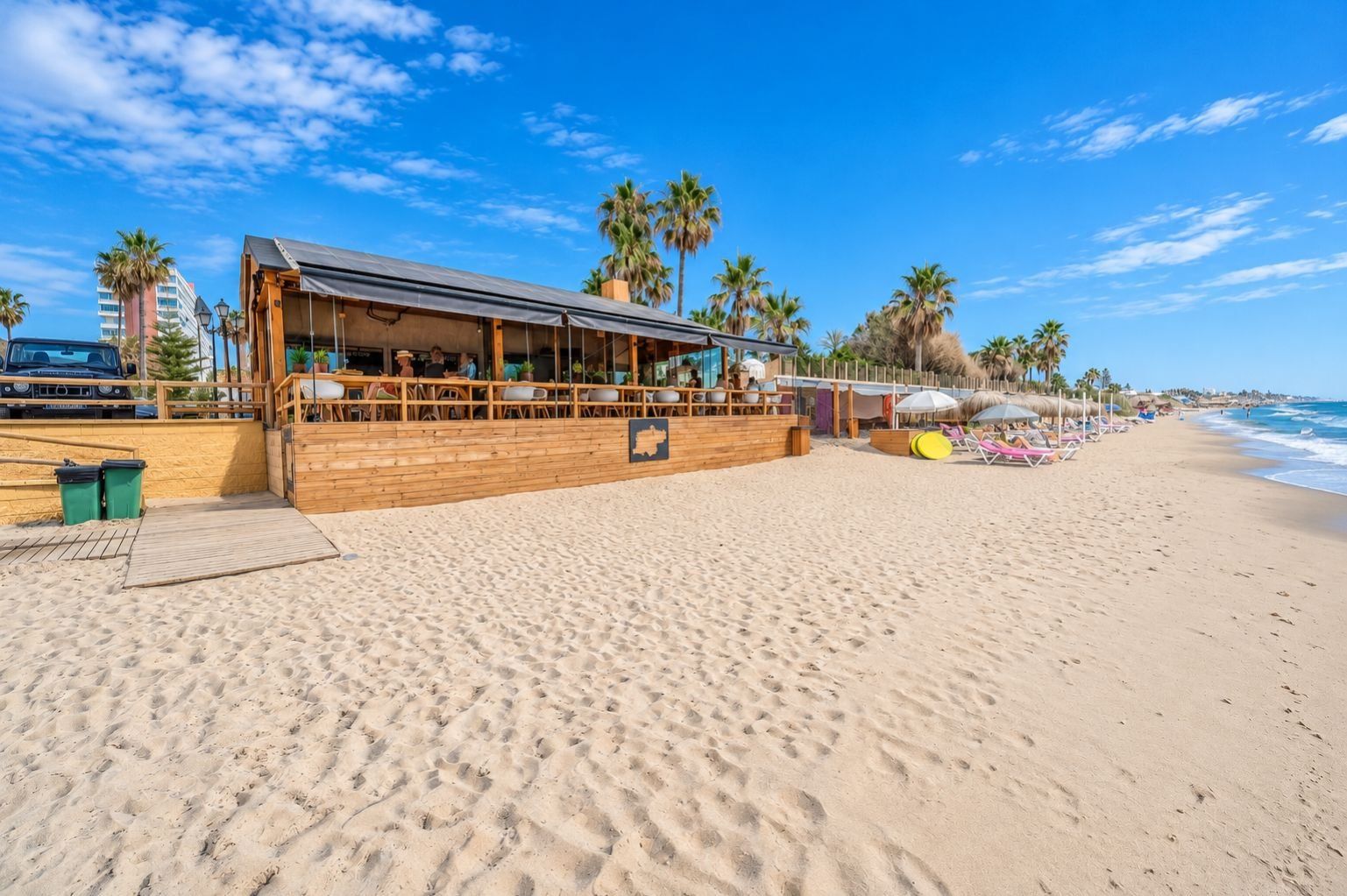 A beachfront restaurant with outdoor seating, palm trees, and a view of the ocean.