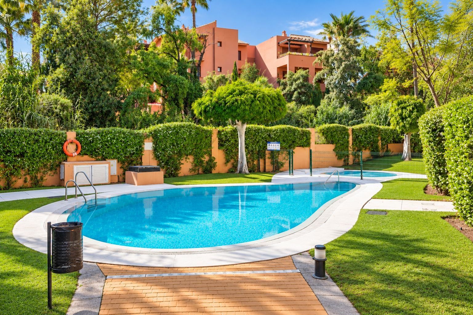 A swimming pool with a wooden deck, surrounded by well-maintained greenery and a building in the background.