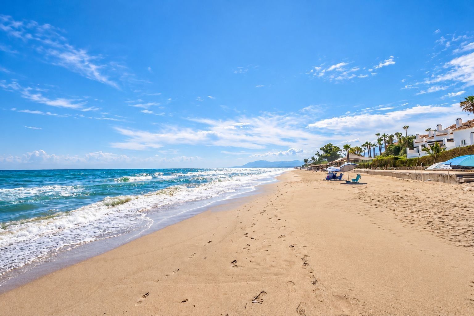 A beach with footprints leading to the water, a row of palm trees, and a building with a sloped roof in the background.