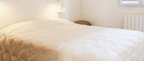 A neatly made bed in a room with a white wall, a window, and a radiator.
