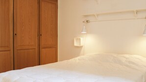 Uma cama com colcha branca, um guarda-roupa de madeira e uma luminária fixada na parede.