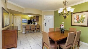 A dining area with a wooden table and wicker chairs, a kitchen in the background, and a green wall with a framed picture. - Westgate resorts  (Orlando)