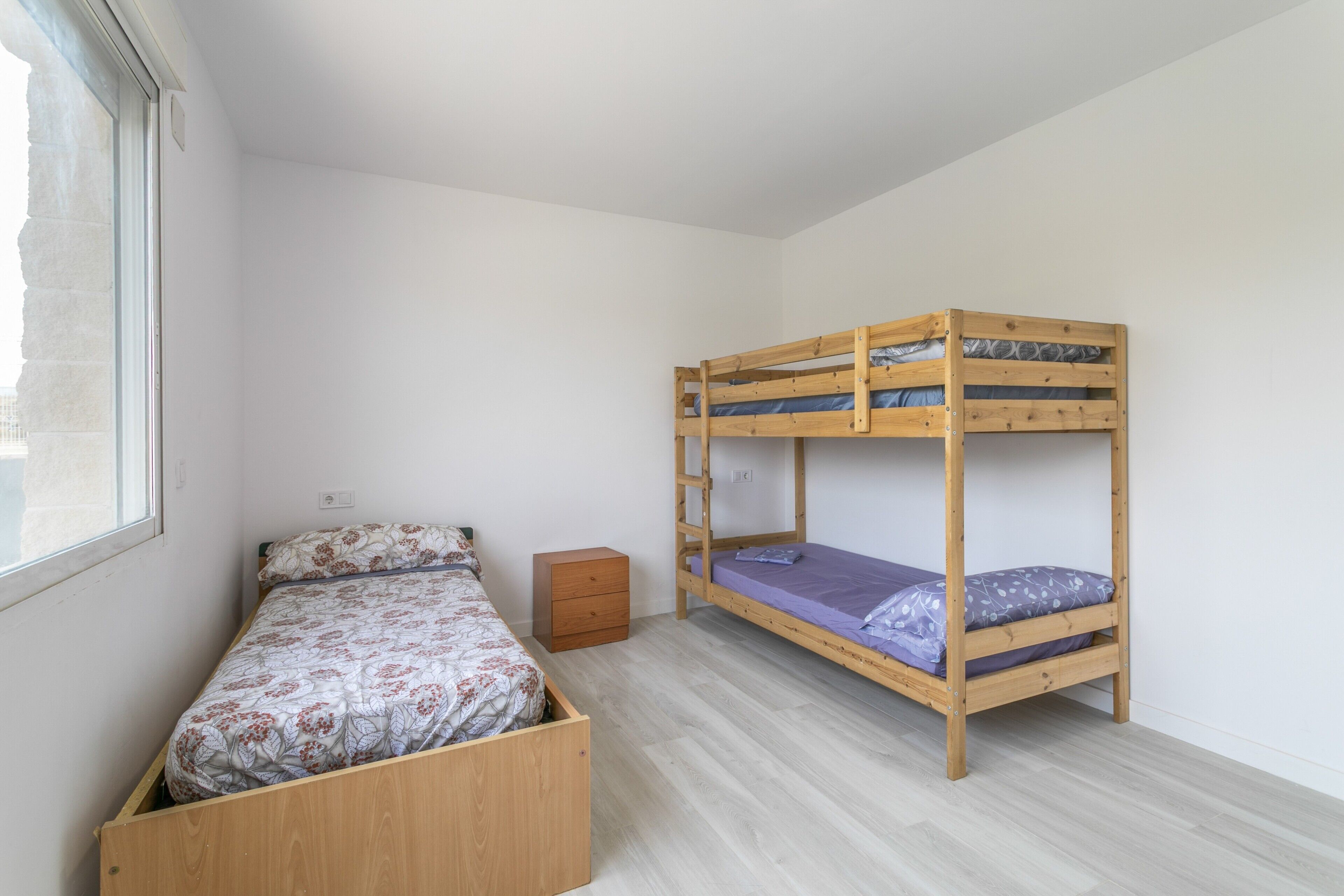 Ein Zimmer mit einem Etagenbett, einem Einzelbett, einem kleinen Holzschrank und einem Fenster.