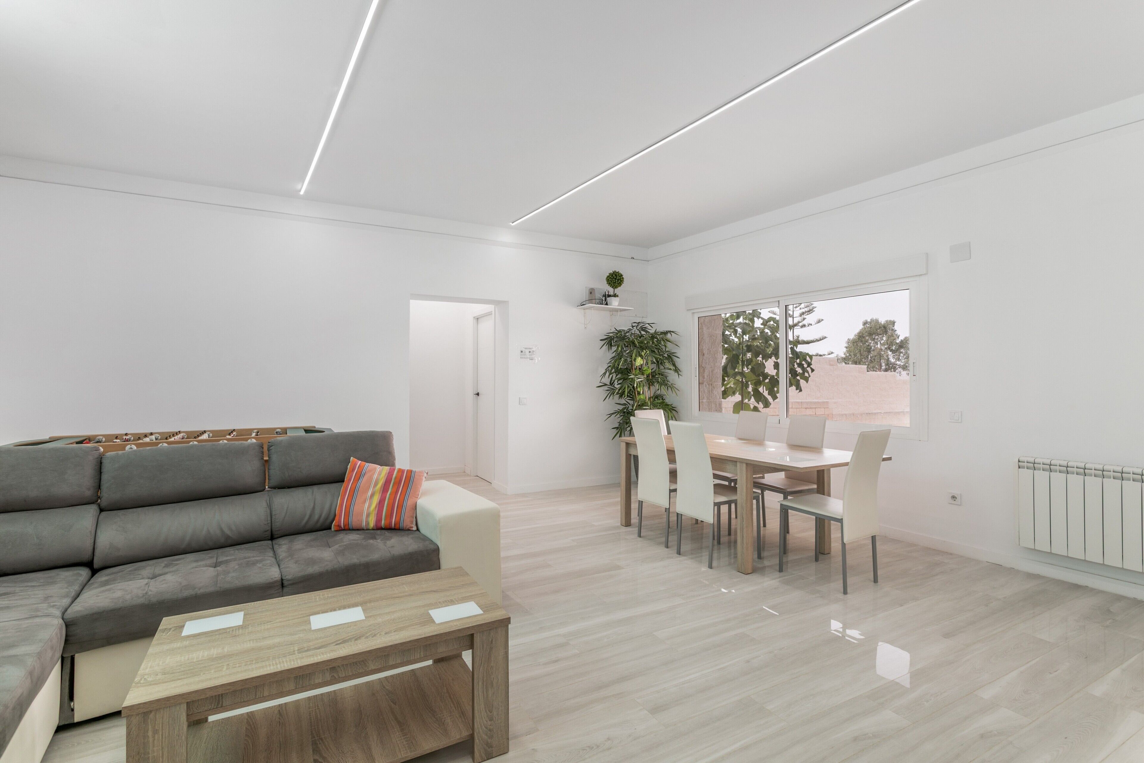 Een moderne woonkamer met een grijze bank, een houten salontafel en een eethoek met een witte tafel en stoelen.