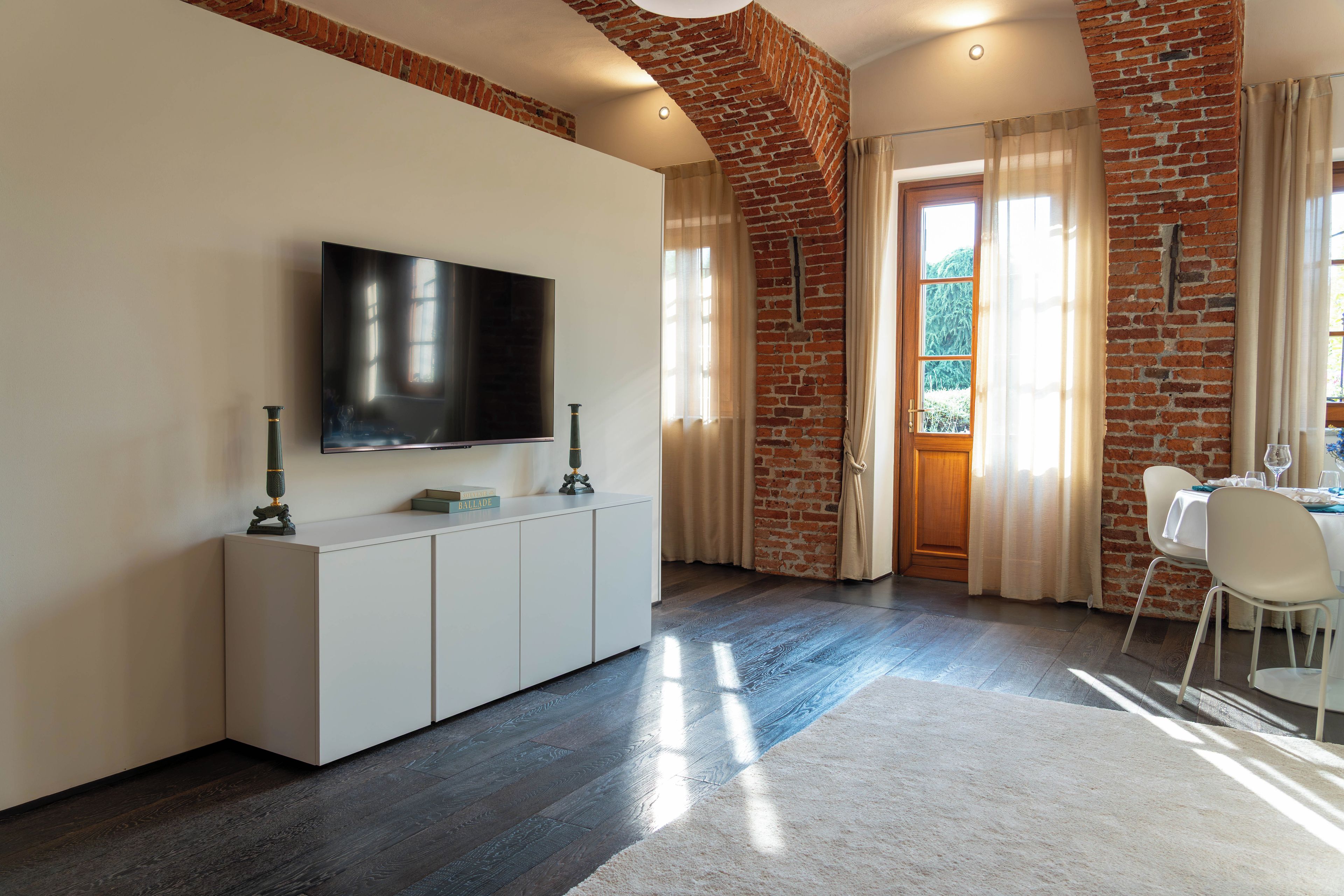 Ein modernes Wohnzimmer mit einem Flachbildfernseher an einer weißen Kommode, einem Essbereich mit Tisch und Stühlen sowie einer Ziegelwand.