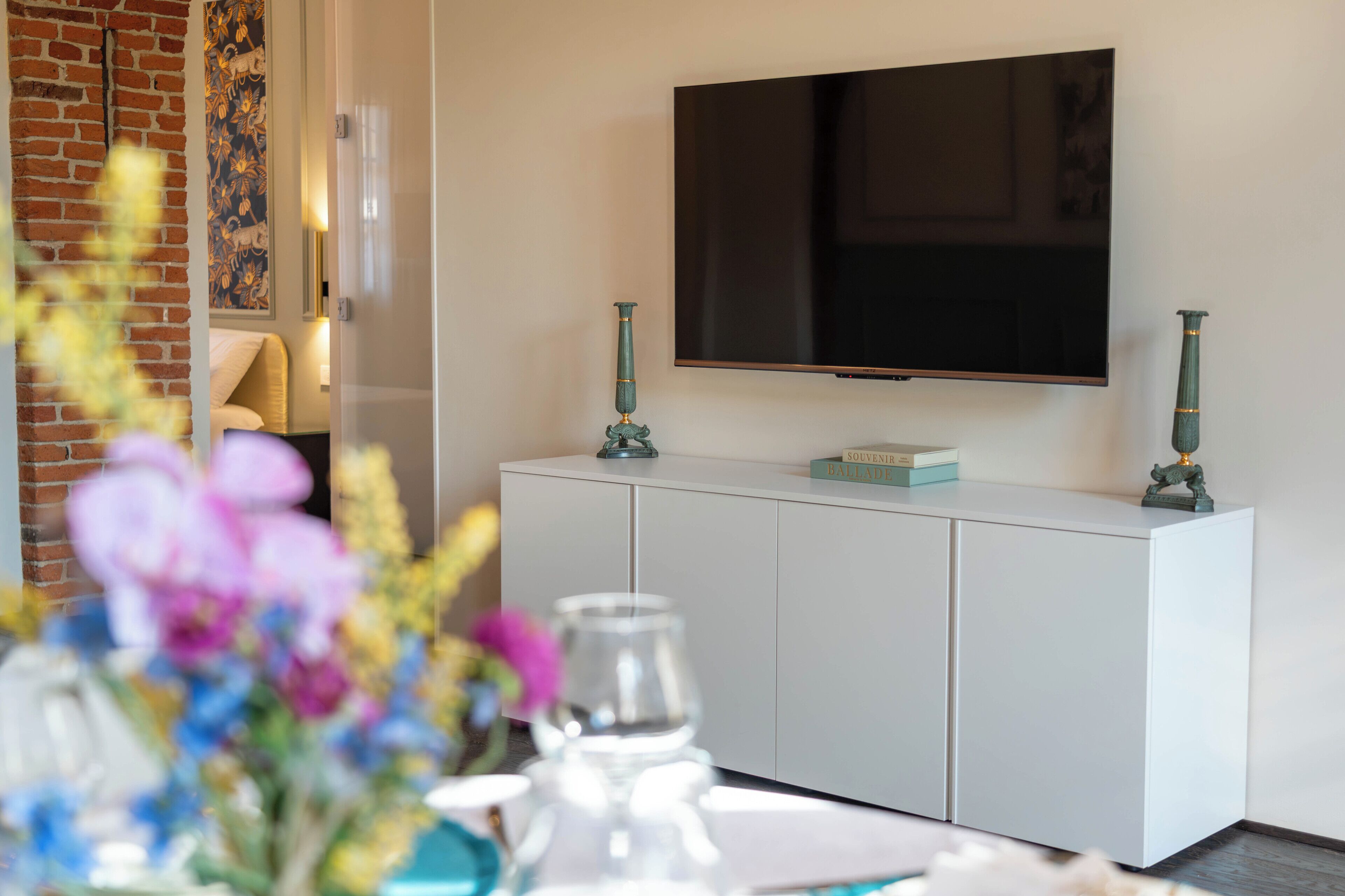 Een moderne woonkamer met een flatscreen televisie aan de muur, een witte kast met decoratieve objecten, en een met bloemen behangen bakstenen muur.