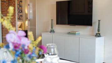 Een moderne woonkamer met een flatscreen televisie aan de muur, een witte kast met decoratieve objecten, en een met bloemen behangen bakstenen muur.