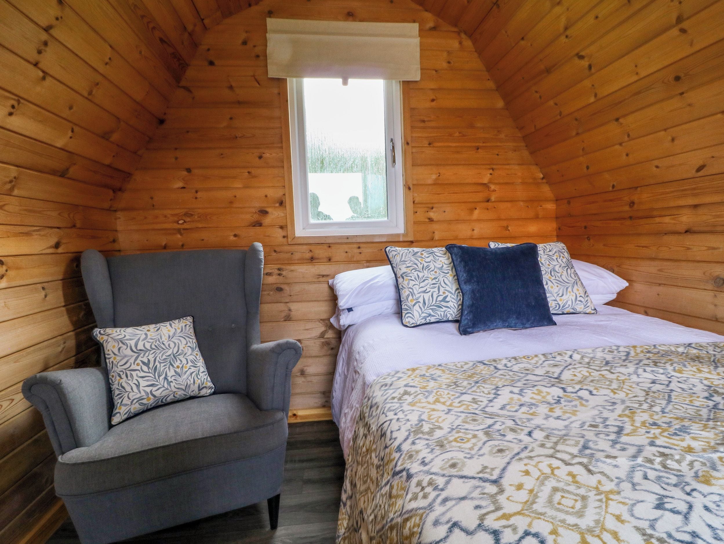Een knus ingerichte kamer met een houten schuine kap, een enkel bed met beddengoed met een patroon, een grijze fauteuil met gekleurde kussens, en een raam met een witte zonwering.