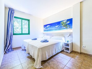 Ein Hotelzimmer mit zwei Betten, einem Fenster mit Blick auf Bäume, einem Strandgemälde und einem gefliesten Boden.