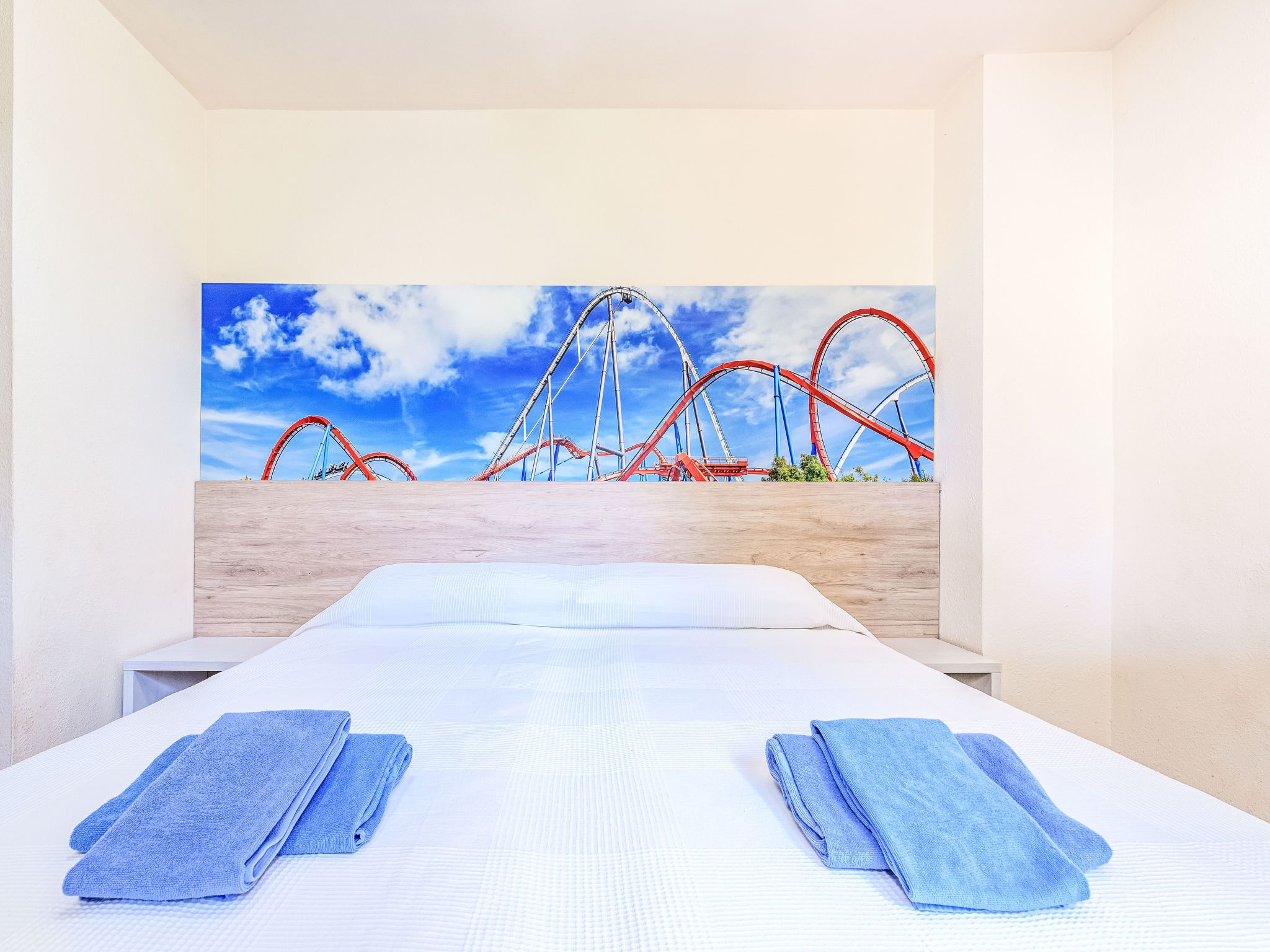 Quarto de hotel com uma cama, duas toalhas azuis e um grande quadro na parede retratando uma montanha-russa.