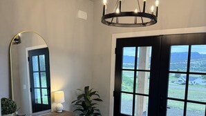 Une pièce avec un plancher en bois, un grand miroir, un lustre, une plante et une vue sur l’extérieur, visible à travers une porte.