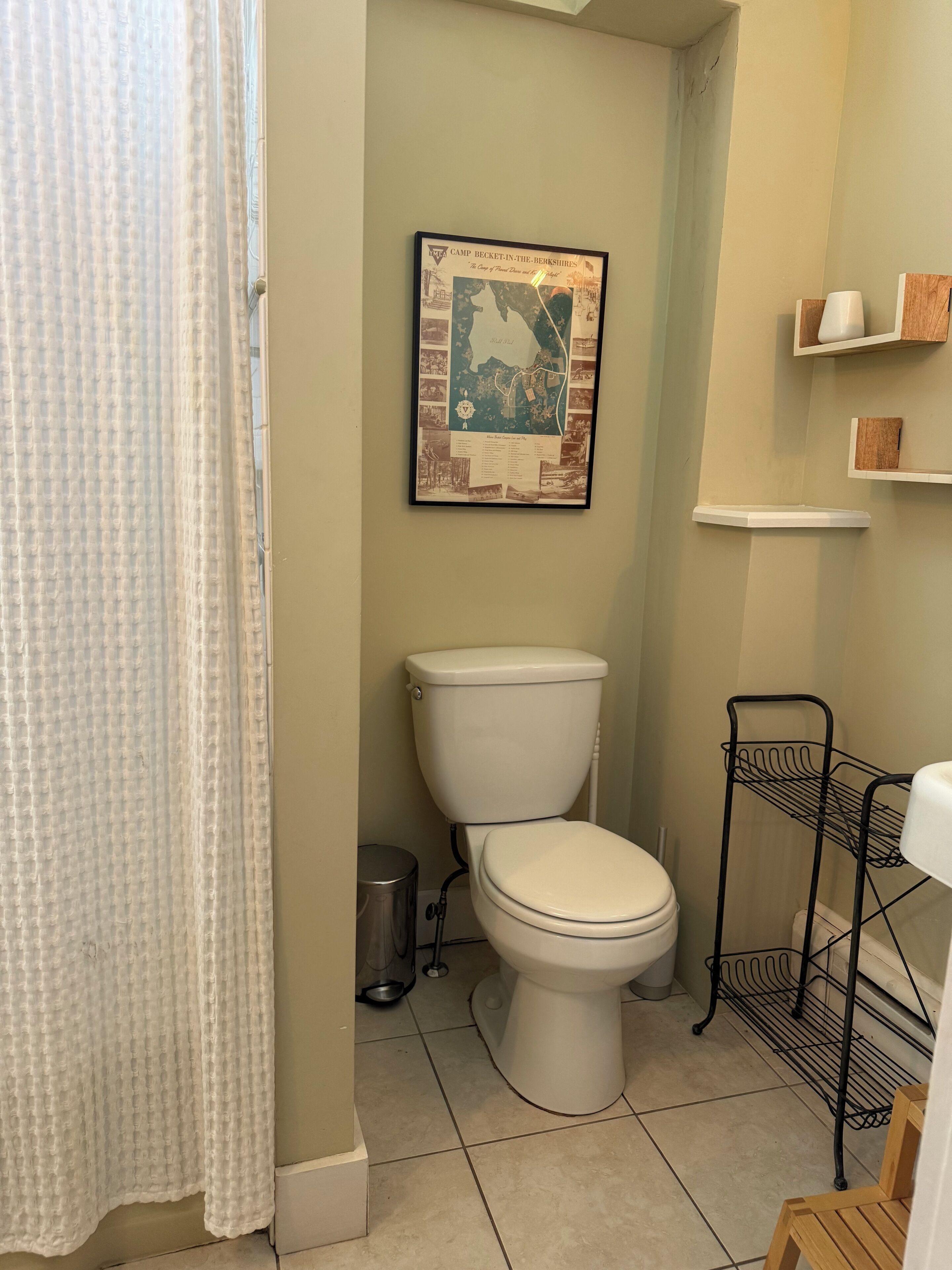 A bathroom with a toilet, a framed map on the wall, a metal rack, and a shower curtain.