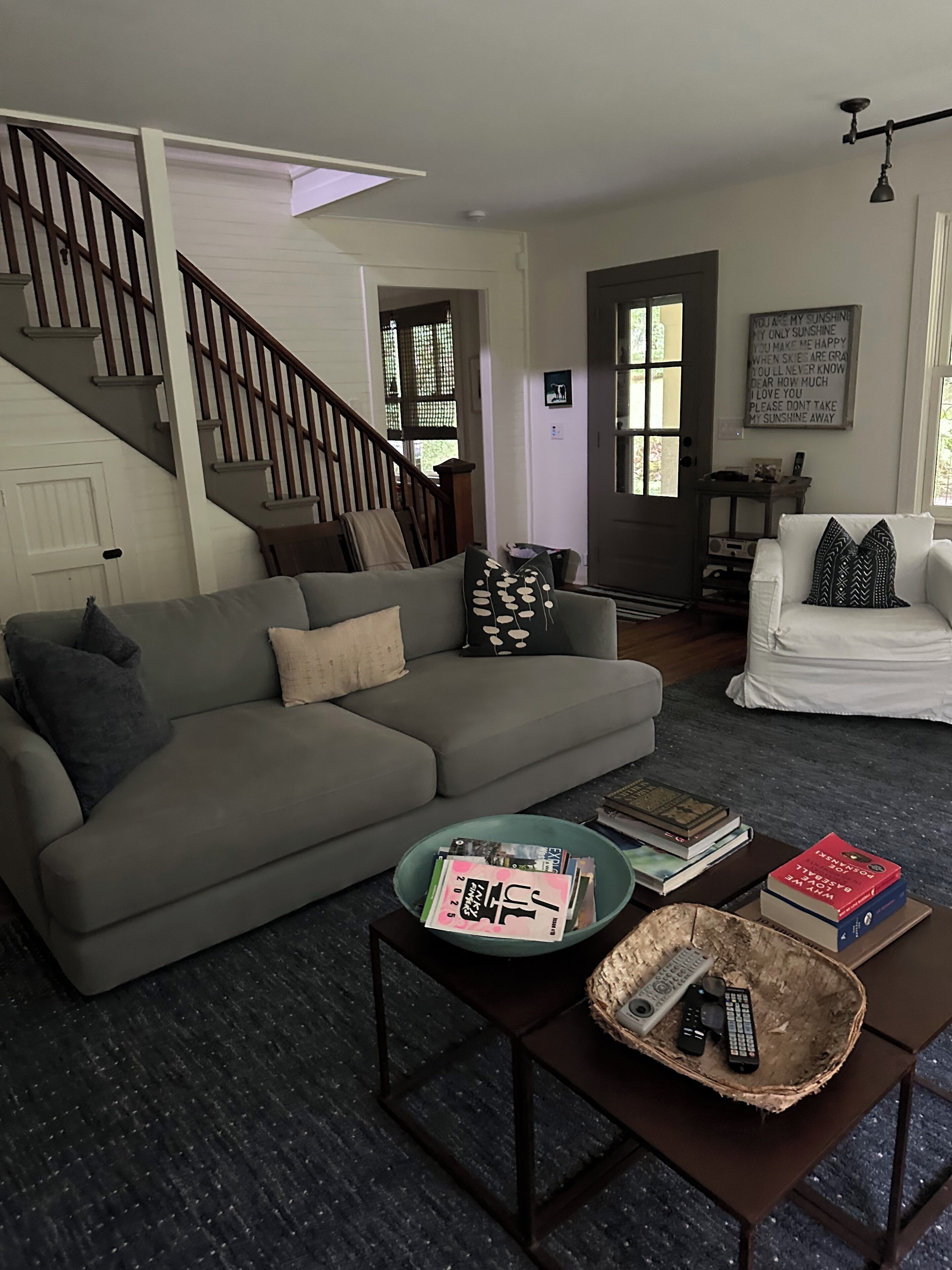 Ein Wohnzimmer mit grauem Sofa, einem Couchtisch mit Zeitschriften und Fernbedienungen, einer Treppe und einer Tür mit einem motivierenden Zitat.