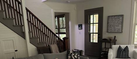 A living room with a grey sofa, a coffee table with magazines and remotes, a staircase, and a door with a motivational quote.
