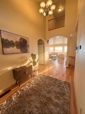 A spacious interior with a high ceiling, a chandelier, a wooden staircase, and a dining area in the background.