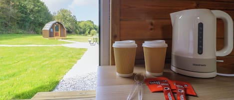 A wooden interior with a view of a green landscape, a small wooden structure, and a white electric kettle on the table.