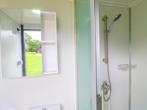 A bathroom with a shower, a mirror, and a view of a green lawn and a building outside. - Skylark Pod (Coalville)