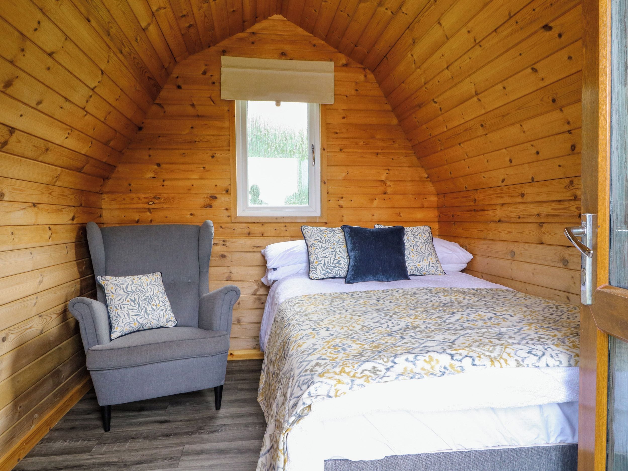 Een gezellige slaapkamer met een schuine kap, een enkel bed met beddengoed met een patroon, een grijze fauteuil met een kussen, en een raam met uitzicht op groen.