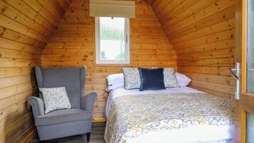 Een gezellige slaapkamer met een schuine kap, een enkel bed met beddengoed met een patroon, een grijze fauteuil met een kussen, en een raam met uitzicht op groen.