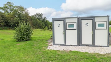 A row of portable restrooms with a small tree beside them, set against a grassy field with trees in the background.