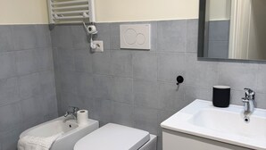 Een badkamer met toilet, bidet, wastafel, spiegel, handdoek en een prullenbak.